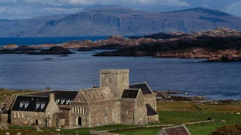 The Abbey at Iona with the sea and rocks in the near distance. In the far distance, some cliffs and hills can he seen. It is sunny.