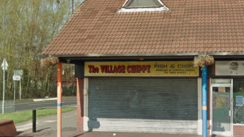 General view of the Village Chippy. It is a unit on a small parade of shops. Its shutters are down in the image.
