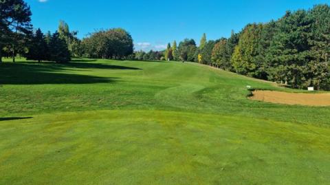 A wide view of a golf course shows a neatly maintained fairway stretching into the distance beneath a clear blue sky. Mature trees line both sides of the hole, creating a natural border of mixed green foliage. In the foreground, a closely mown green leads towards the fairway, while a sand bunker is visible on the right-hand side. It is a sunny day with a blue sky.