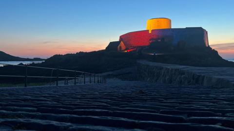 A view of Fort Grey lit up in red, white and blue. The sun is setting in the background.