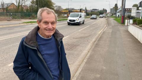 Roy Egan, pictured smiling next to the road. He has grey hair and wears a blue jumper and a darker blue jacket. The road is wide, and a petrol station is visible in the background. Traffic, including a white van, is seen travelling towards the camera. 