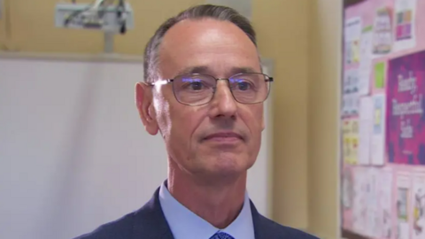 Mark Browne  has receding grey/brown hair and brown eyes and glasses standing in front of a a school white board and pink billboard. He is wearing a navy suit, white tie and blue shirt and has a neutral expression.