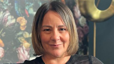 A head shot of Sue Greenwood. She has mid-length blonde/brown hair and is sitting on a sofa in front of green floral wallpaper. She is wearing a long sleeve black top and a gold necklace. 