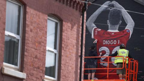 Two people on a scissor lift spray-painting the final touches to a mural of Diogo Jota on the side of a house