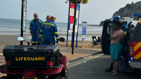 A man wearing green shorts smokes a cigarette as he stands near lifeguards wearing blue uniforms and lifeguard vehicles. A sandy beach is in the background.