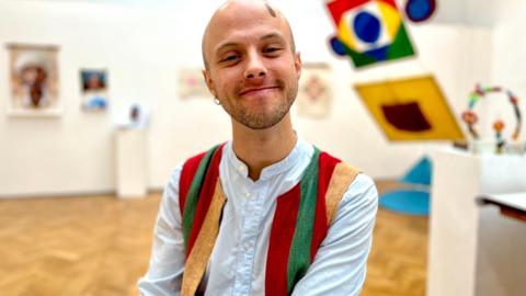 Brook wearing a pale blue shirt and colourful waistcoat in a gallery room smiling at the camera