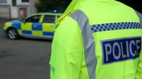 Police officer, photographed from behind, wears a hi-vis jacket. A police patrol vehicle is behind the officer.