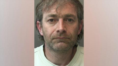 A police head and shoulders shot of a man with grey and brown hair that isn't styled and is nearly at his shoulders. He has a short stubbled moustache and beard and is wearing a white top and looking directly into the camera. 