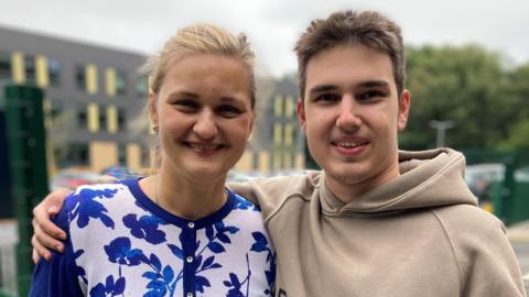 A woman in a blue and white floral shirt smiling with her arm round her son, who is wearing a beige hoody 