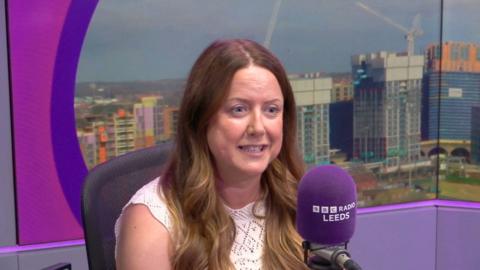 A woman with long brown curly hair wears a white lace top. She is smiling and sat in front of a purple microphone