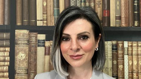 Lucy Cohen smiles at the camera in front of a bookshelf. She has brown eyes, dark hair with blonde streaks and a septum piercing 