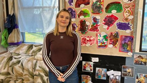 Stacie Moxon with blonde, shoulder-length wearing a brown top and blue jeans. She is standing in front of a wall with pictures drawn by the children