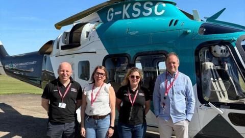 Duncan Bootland, Sophy Dunn, Helene Poursain and Warren Porritt smiling for the camera in the sun. They are stood beside a blue and white KSS helicopter.