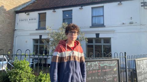 Emma looks into the camera – she wears a hooded jacket in red, white, brown and blue. Behind her is a whitewashed pub building, with chalkboards outside with menus and offers. The sign Coates House is painted onto the building.