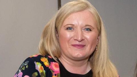 A woman with blond hair and a bright multi-coloured top on, smiling at the camera 