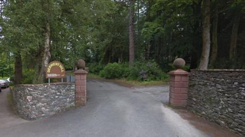 The entrance to Derwent Hill outdoor centre. A sign with the centre's name is displayed on a stone wall. An asphalt road leads to the centre through woodland. Trees line either side of the road.