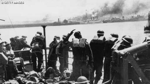 Troops stand on a pier and look out at the sea at Dunkirk. In the distance there are smoke and ships.