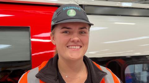 A woman is wearing a black Siemens' t-shirt with an orange hi-vis vest on and with a black Siemens' cap. She is smiling at the camera in front of a tube carriage that she is working on.