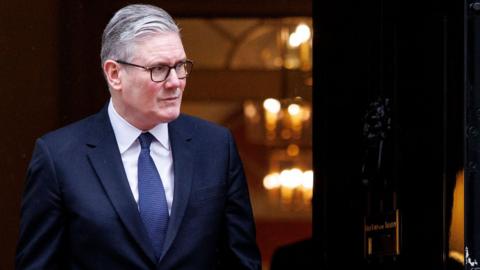 Prime Minister Sir Keir Starmer standing in front of an open door to 10 Downing Street in London, in a navy suit and blue tie looking to the right.