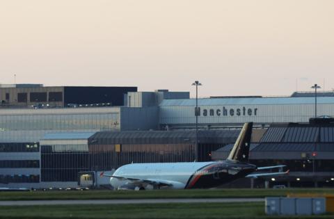 An airplane is on the runway, with grass in the foreground and an airport in the background with Manchester emblazoned on it