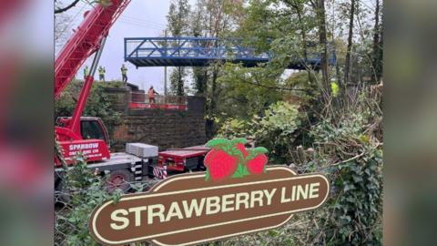 A bridge being lifted on a crane over a road. In the foreground is a sign for the Strawberry Line.
