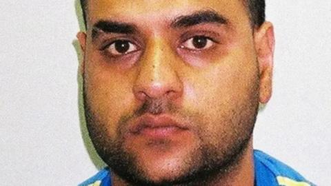 A mugshot of Mohammed Ali Sultan looking at the camera. He has a beard and a slash in his left eyebrow. He is wearing a blue t shirt with yellow stripes on the shoulder