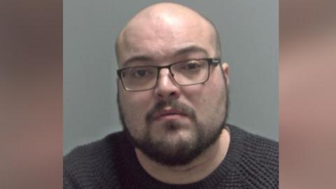 Geddes Abbott a man who is looking directly at the camera and is wearing a dark grey sweater and glasses.