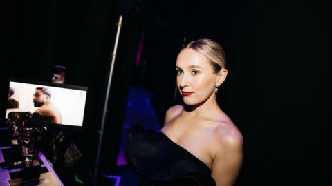 A glamorous woman in a black dress stands in the darkness behind a stage. 