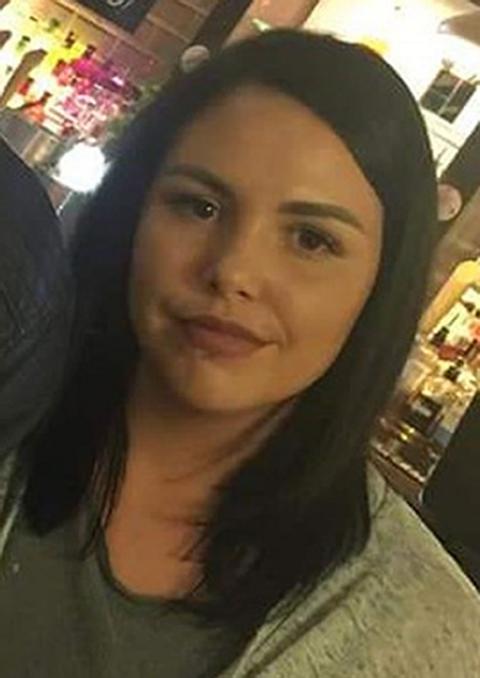 A young woman with shoulder length dark brown hair smiles at the camera. She is indoors, in a dimly-lit room