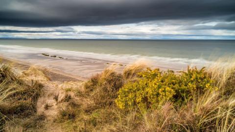 A view over sand dunes towards the beach at Findhorn. The tide is out and there is a stretch of sand along the water's edge. There are dark grey clouds overhead.