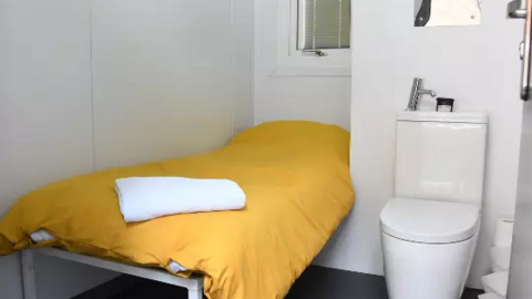 The interior of a homeless pod, which shows a small white room with a bed with a yellow duvet and a toilet next to it.