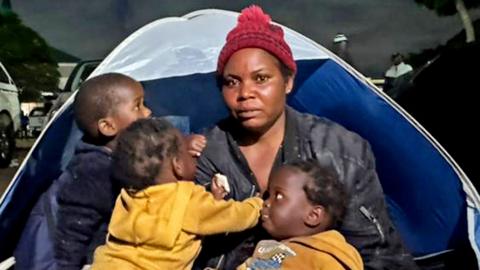 Esnat Joseph, wearing a maroon woollen bobble hat and black jacket, sitting in front of a tent with her baby triplets - two dressed in yellow hoodies and one black.