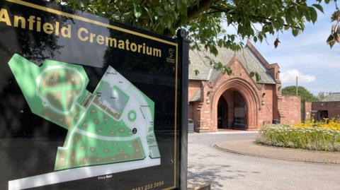 A black sign with gold writing which says Anfield Crematorium in front of a red building set behind trees with a flower bed in front.