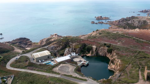 An aerial view of the desalination plant off the coast. A road can be seen winding down towards a large building with cars parked outside it and a body of water. Rocky surroundings face the ocean.