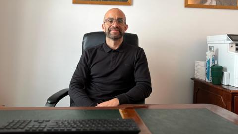 Jason looks at the camera as he he is sat behind a desk. He is wearing a black jumpers with a collar and glasses. Jason has stubbly facial hair and his arms are slightly crossed. There is a keyboard and mouse in front of him and a printer on a cabinet to his right. 