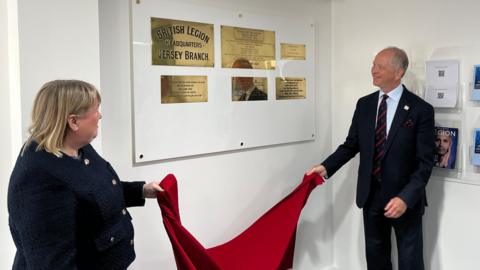 The image shows two people holding some red fabric unveiling a series of commemorative brass plaques. They are in a room with white painted walls.