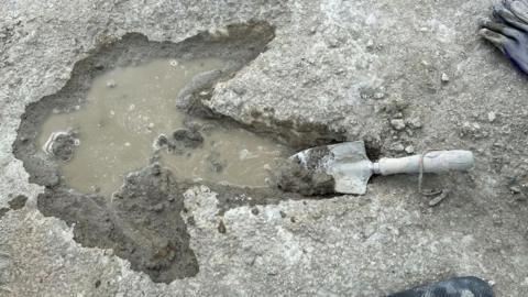 A single track dinosaur footprint. The large print is full of greyish water with a trowel in one of the three toes. The print is in a grey stone. There is a black glove in one corner of the photo.