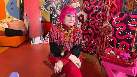 Zandra Rhodes with short pink hair sitting on a red chair wearing red trousers and a red shirt. She is in a colourful room which is mainly red and pink.