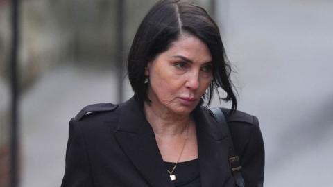 Sadie Frost, a woman with short, straight black hair, looks down with serious expression. She is wearing a black jacket above a black top, with a gold necklace.