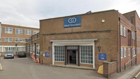 The front entrance to a manufacturing site - an old brick building. There are windows either side of the open front door. A sign above says Ideal Standard while signs either side of the entrance say Ideal Standard, Armitage Shanks.