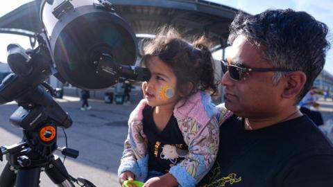 Tawhid Rana, of Midland, Michigan., hold his daughter Thia, as she views the sun through a telescope at the Indianapolis Motor Speedway in Indianapolis, Monday, April 8, 2024.