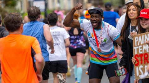 A person wearing colourful cycling gear and a dark cap cheering on runners surrounded by a crowd of people