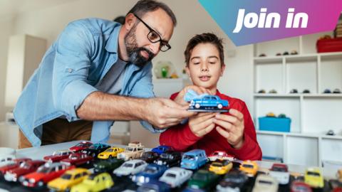 A boy and his dad looking at a toy car which makes up part of a toy car collection laid on the table in front of them.