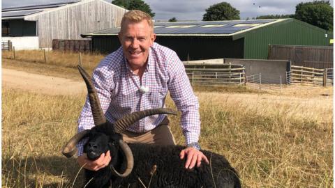 Adam Henson at Cotswold Farm Park, posing with a goat in front of a barn with solar panels