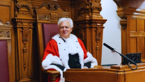 Robert MacRae in a chamber. He is sitting on a wooden seat which is lined with leather and modeled a little like a throne. He is smiling at the camera and is wearing a red robe with white ermine trim over a black suit.