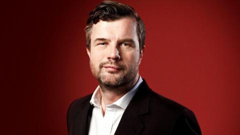 BBC Radio 3 presenter Petroc Trelawny has dark hair brushed over to one side and designer stubble. He is wearing a black suit jacket over a white shirt. He is photographed against a red background.