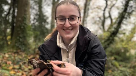 Niamh Carmichael, a woman with fair hair, glasses, dark jacket and brown fleece, holding leaves in her hands.