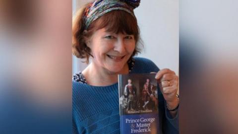 Rosalind Freeborn is wearing a blue jumper and is smiling at the camera. She is holding a copy of her book, Prince George and Master Frederick.