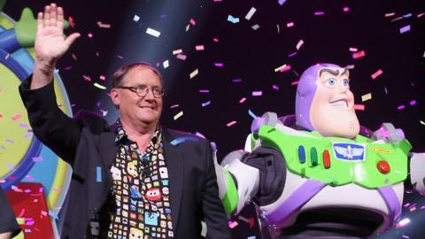 John Lasseter waves to a crowd from the stage next to a costumed performer appearing as popular Toy Story character Buzz Lightyear