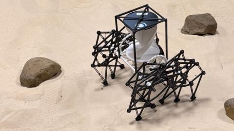 A robotic creation comprising a black plastic cuboid frame, raised off the ground by two black plastic geometric shapes attached by white plastic cogs. The structure is pictured on sand, beside three small brown rocks 